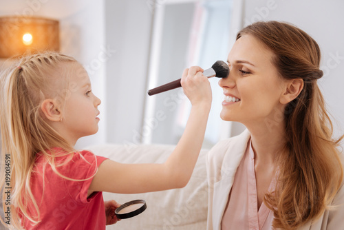 Facial makeup. Cheerful pleasant blonde girl holding a brush and applying powder while doing make up for her mother