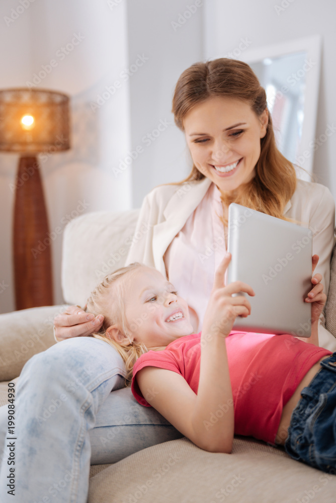 Digital technology. Cheerful delighted young girl lying on her mothers laps and looking at the tablet screen while resting at home Digital technology. Cheerful delighted young girl lying on her mothers laps and looking at the tablet screen while resting at home