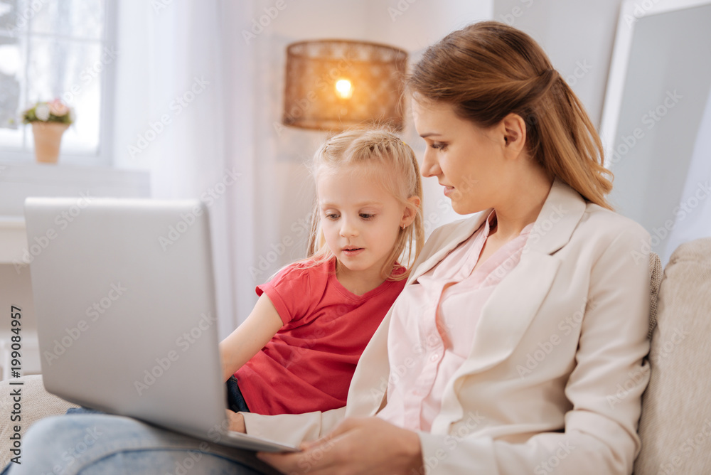 Modern device. Nice pleasant young woman holding a laptop and looking at her daughter while being at home with her