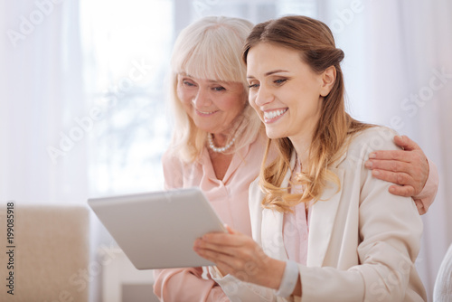 Digital gadget. Delighted positive young woman smiling and looking at the tablet screen while sitting together with her mother