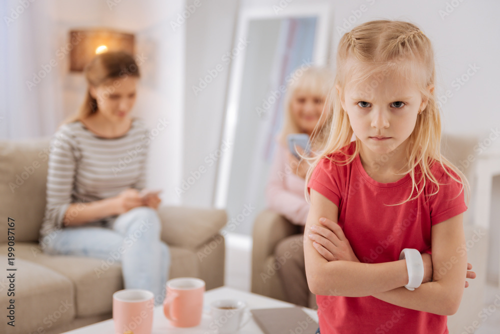 Emotional child. Unhappy cheerless blonde girl sitting on the sofa and turning away from her mother and grandmother while being in a bad mood Emotional child. Unhappy cheerless blonde girl sitting on the sofa and turning away from her mother and grandmother while being in a bad mood