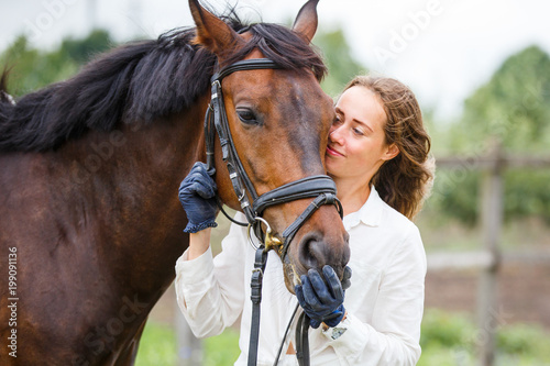 Young happy rider woman with her bay horse. Portrait of smiling girl with her stallion in paddock on summer day