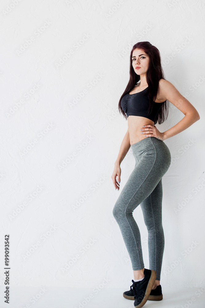 Full-length side portrait of the young athletic woman in black short top and gray leggings posing at white background. Full-length side portrait of the young athletic woman in black short top and gray leggings posing at white background.