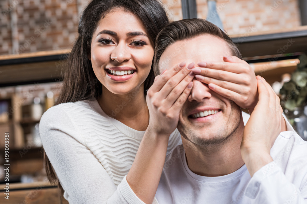 happy young african american woman closing eyes to smiling caucasian boyfriend at home happy young african american woman closing eyes to smiling caucasian boyfriend at home