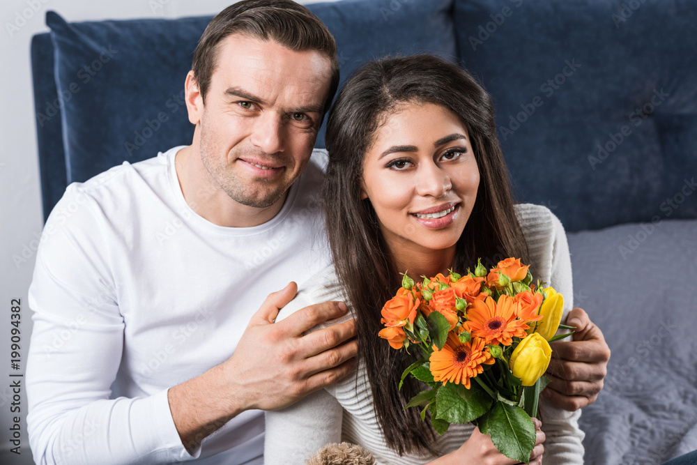 happy young man embracing girlfriend with bouquet of flowers and smiling at camera