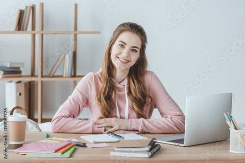 smiling female student sitting at table with laptop and copybooks