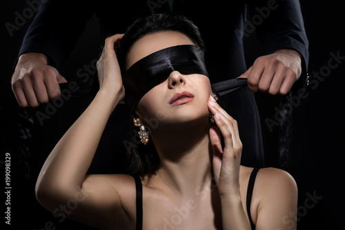 Man in suit ties a woman's eyes with a silk ribbon on black background