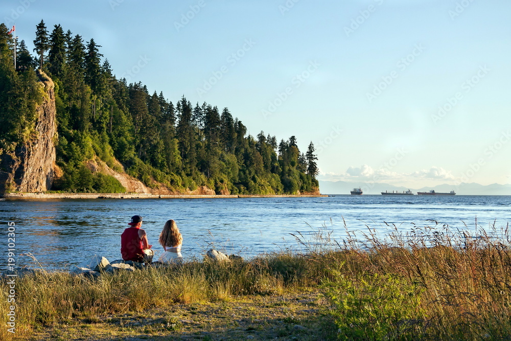 A young couple sits at the shore of the bay, on the opposite shore a wooded cliff, blue sky, several merchant ships on the horizon. A young couple sits at the shore of the bay, on the opposite shore a wooded cliff, blue sky, several merchant ships on the horizon.