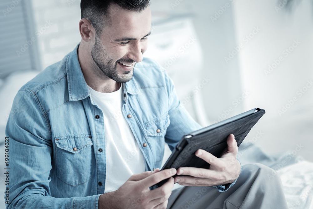Modern technology. Delighted positive handsome man holding a tablet and smiling while using it