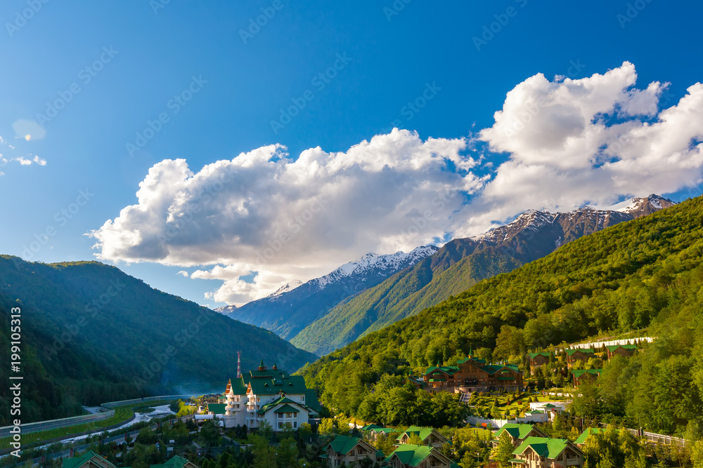 Modern mountain cottages with green roofs against the backdrop of a mountain valley covered with a green spring sun forest. Ski Resort at Caucasus Mountains, Krasnaya Polyana, Sochi, Russia.