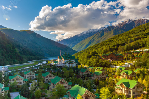 Modern mountain cottages with green roofs against the backdrop of a mountain valley covered with a green spring sun forest. Ski Resort at Caucasus Mountains, Krasnaya Polyana, Sochi, Russia. photo