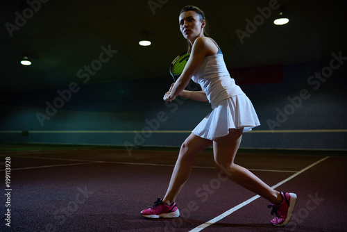 Full length motivational portrait of beautiful young woman playing tennis in dark indoor court, ready to hit ball, copy space