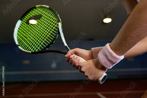 Close up of unrecognizable woman holding racket while playing tennis in indoor court and swinging to hit ball, copy space