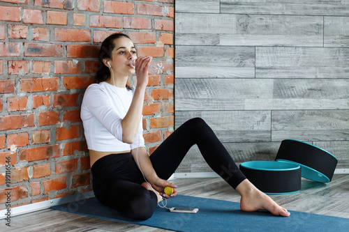 Tired young woman sits on the floor, listens to the music and drinks water after work out in gym