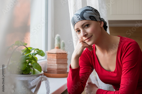 Young positive adult female cancer patient sitting in the kitchen by a window, smiling and looking at the camera.