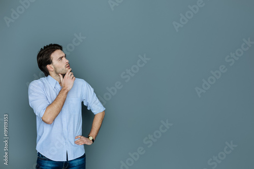 Thoughtful look. Smart handsome thoughtful man holding his chin and looking up while standing against the grey background