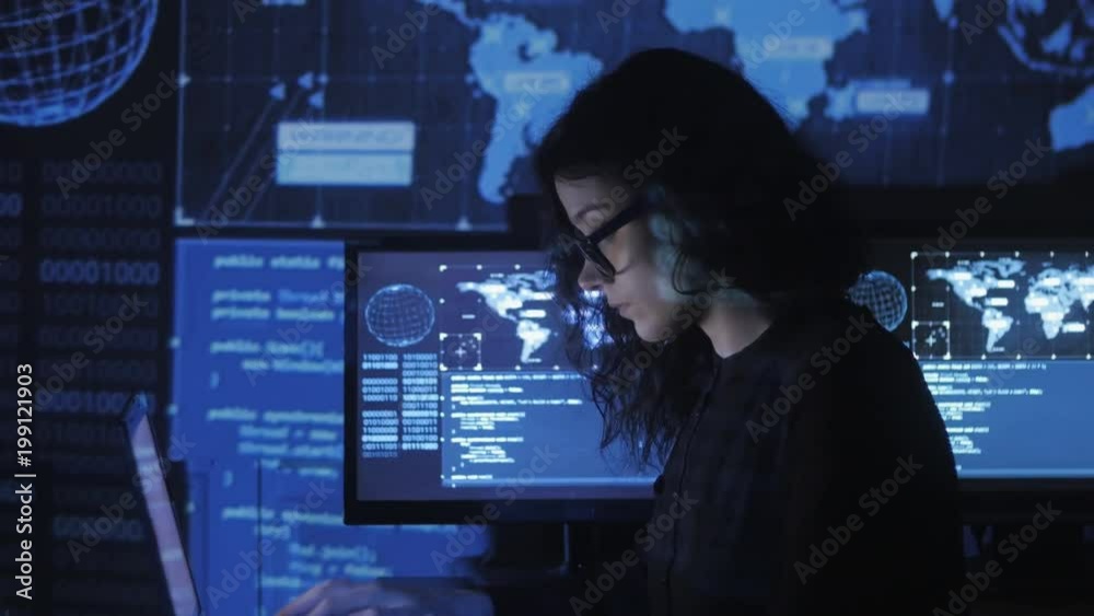portrait of young woman programmer in eyeglasses working at a computer in the data center filled with display screens