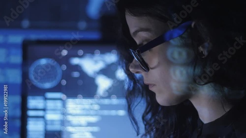 portrait of young woman programmer in eyeglasses working at a computer in the data center filled with display screens