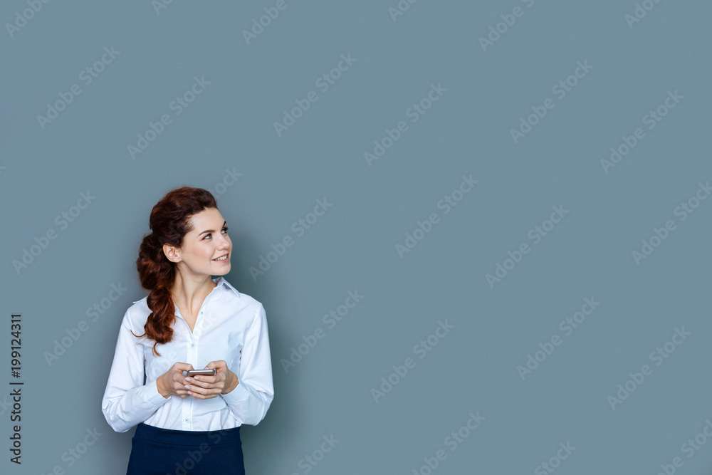Career ladder. Joyful nice smart woman smiling and holing her smartphone while working in the office Career ladder. Joyful nice smart woman smiling and holing her smartphone while working in the office