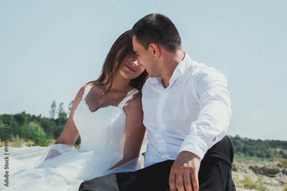 Wedding couple is sitting and hugging on the white sandy lake beach. Romantic hug and embrace of bride and groom in summer heat love story. Heating sun and weather. Travel lifestyle photo.