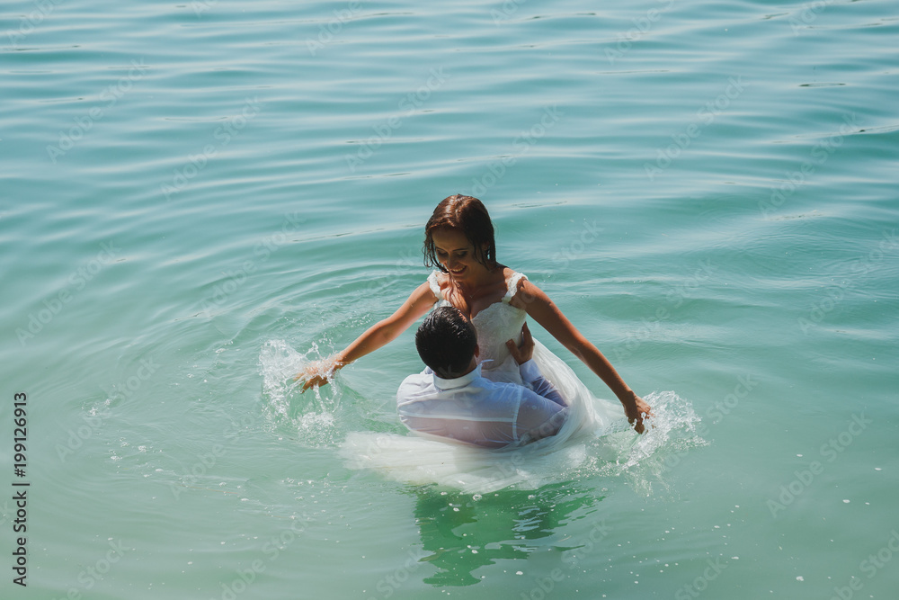 Wedding couple is hugging in azure blue lake water. Beautiful bride in puffy dress and groom are having fun. Summer passion crazy emotions photo on the seaside. Wet wedding clothes.