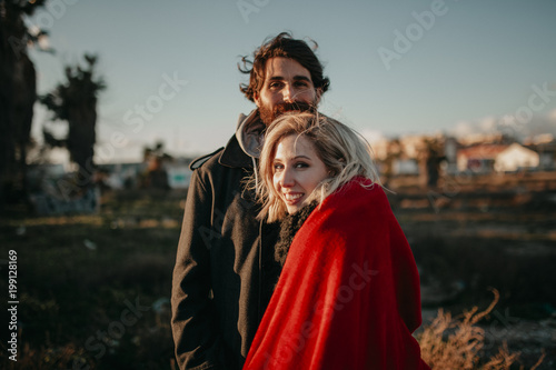 Close up portrait of young alternative couple, outdoors, at sunset, while they look at the camera smiling.
