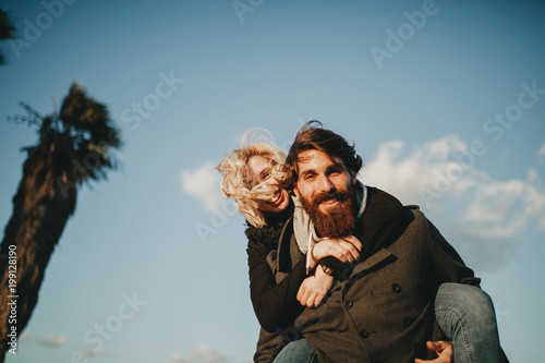 Cool indie couple having fun outdoors while he gives her a piggyback, while they look at camera.