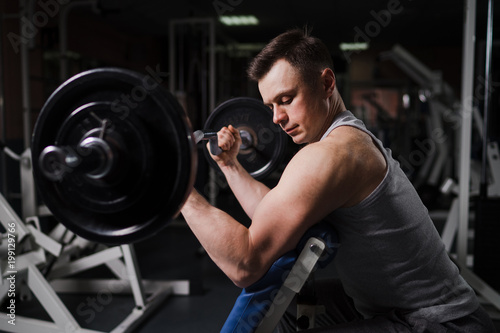 Strong handsome fit man exercising in the gym. Personal trainer workout. Athletic man working out his arms muscles with barbell on a bench. Fitness, healhty lifestyle, bodybuilding concept.