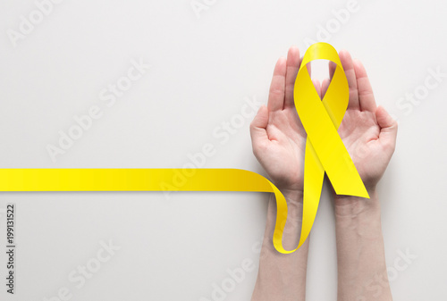 Two hands holding yellow ribbon, symbol of cancer awareness, medical support and prevention with helping hand. Place for text.
