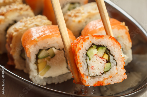 Two chopsticks holding Uramaki California and different  sushi rolls with seafood on ceramic plate on the background