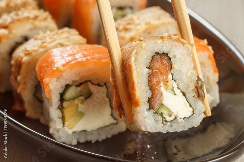 Two chopsticks holding Uramaki with Conger and different sushi rolls with seafood on ceramic plate on the background