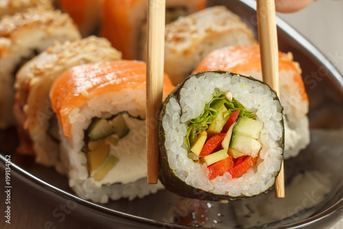 Two chopsticks holding Hosomaki with vegetables and different sushi rolls with seafood on ceramic plate on the background