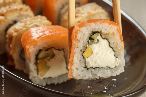 Two chopsticks holding Uramaki Philadelphia and different sushi rolls with seafood on ceramic plate on the background