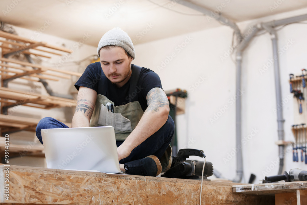 a man in working clothes is typing in a laptop. Portrait of man at work in workshop in garage at home a man in working clothes is typing in a laptop. Portrait of man at work in workshop in garage at home