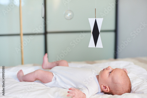 Adorable baby boy in white sunny bedroom lying and looks at Munari Montessori mobile. photo