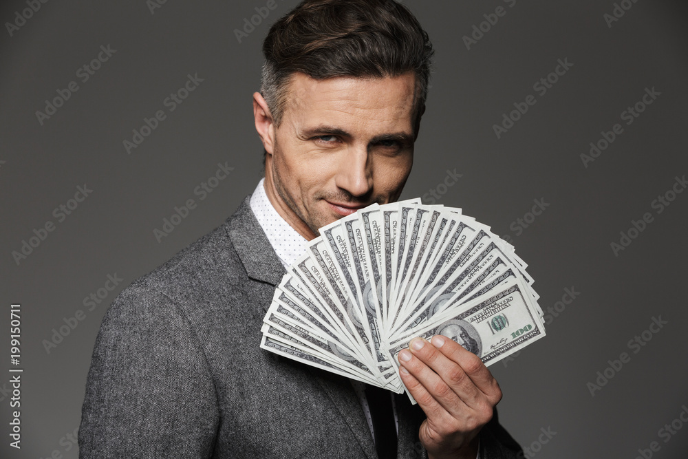 Photo of rich and happy entrepreneur guy 30s in business suit demonstrating money prize in dollar cash and looking on camera, isolated over gray background Photo of rich and happy entrepreneur guy 30s in business suit demonstrating money prize in dollar cash and looking on camera, isolated over gray background
