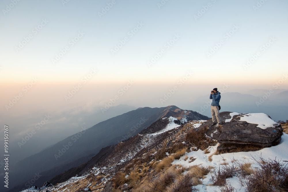 Hiker staying on snow mountain top above clouds and valley, doing pictures on small photo camera