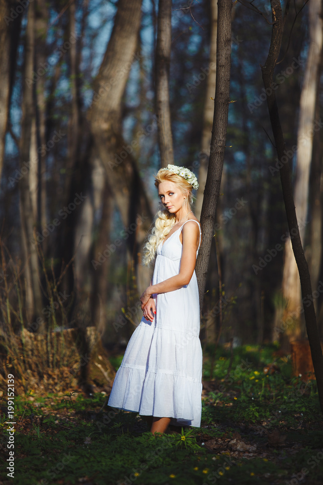 blond woman stands in white dress by thin tree.