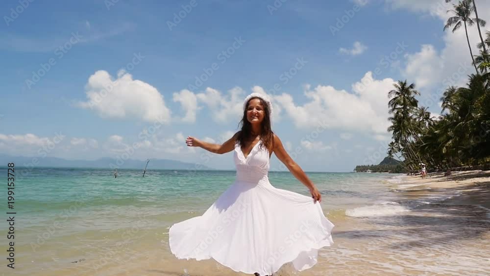Pretty young woman in a white dress and flower wreath runs along the seacoast barefoot and enjoys herself. Beach with white sand and the sky with clouds. Slow Motion. 1920x1080