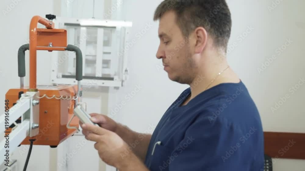 Adult male doctor in blue uniform adjusts using an X-ray control panel. Assistant sets up medical tools. Adult male doctor in blue uniform adjusts using an X-ray control panel. Assistant sets up medical tools.