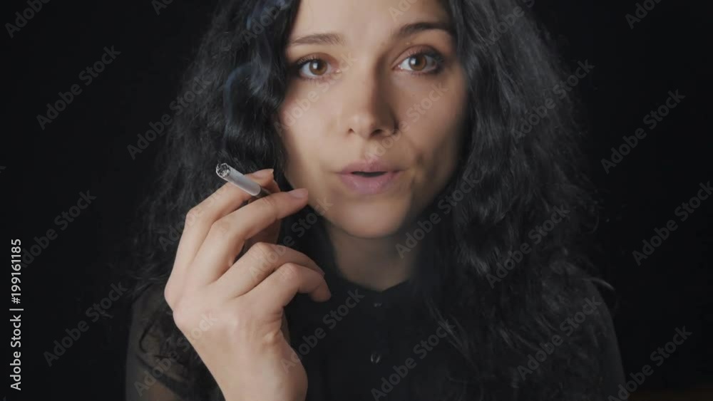 Portrait of young brunette woman with curly hair is smoking a cigarette and looking at the camera on the dark background