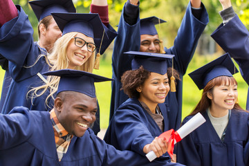education, graduation and people concept - group of happy international students in mortar boards and bachelor gowns with diplomas celebrating success
