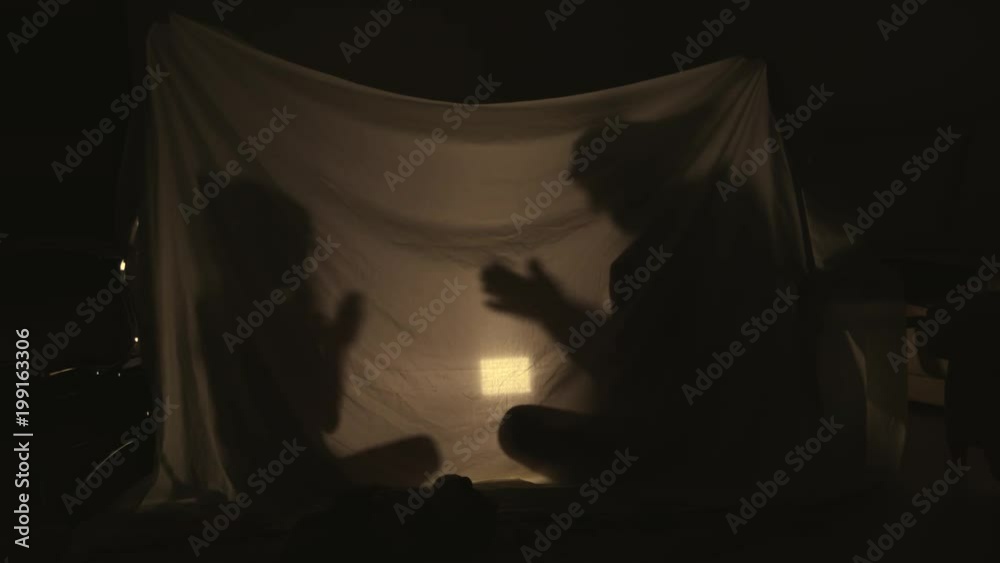 Silhouette of mom and daughter playing together inside a blanket fort in the evening, lit by a lamp from inside.