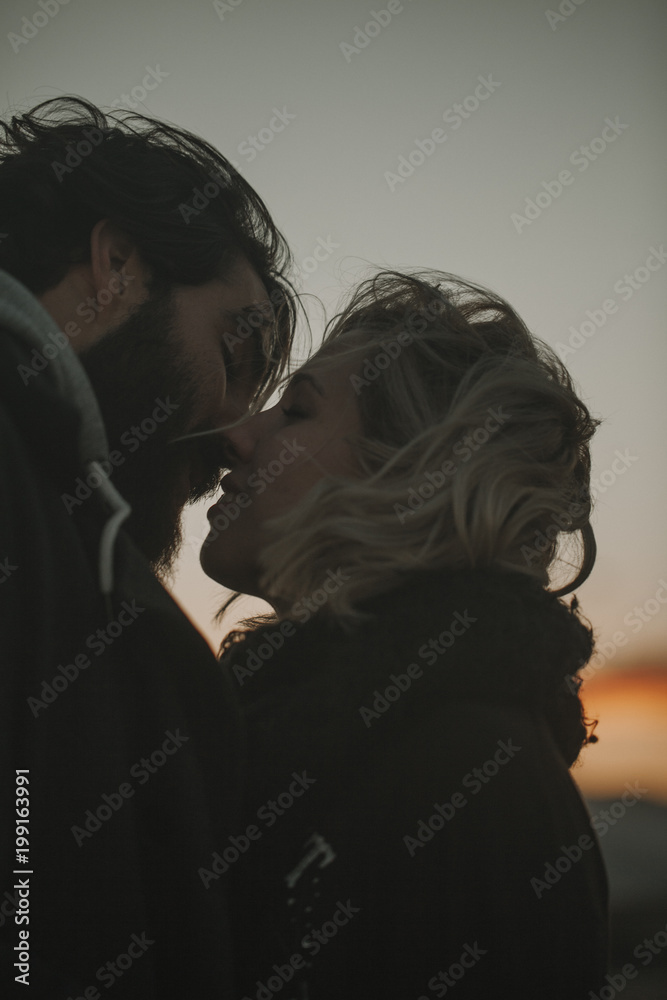 Close up portrait of cool couple kissing at sunset with an orange sky in the background.