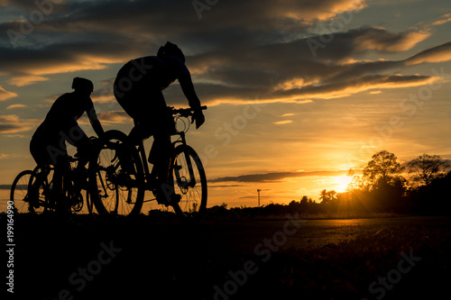 The men ride bikes at sunset with orange-blue sky background. Abstract Silhouette background concept.