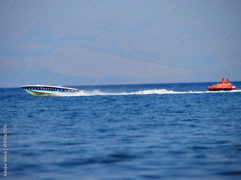Naklejka premium Water amusement on banana boat. summer holiday by the sea. Beautiful bright blue water and red and white boat and yellow banana boat.
