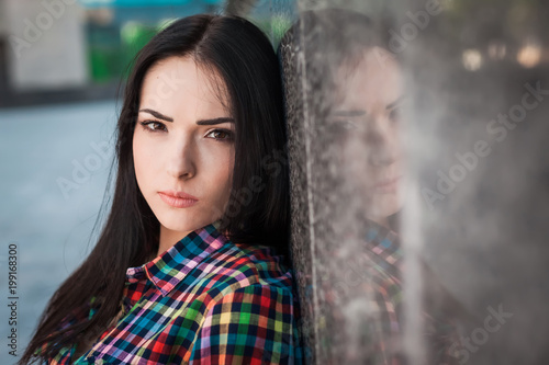 Portrait of young hipster teenager with long hair leaning against the wall in the street, wearing colorful shirt in cage on the urban background