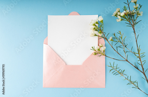 Minimal composition with a pink envelope, white blank card and a wax flower on a blue background. Mockup with envelope and blank card. Flat lay. Top view.