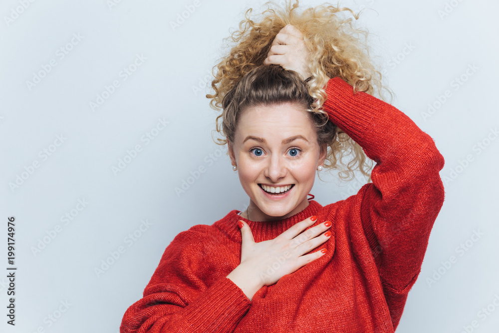 Adorable Excited Young Female With Curly Hair Keeps Hair Tied In Pony Tail Dressed In Casual Red Sweater Happy To Recive Positive News From Interlocutor Poses Against White Studio Background Stock Photo