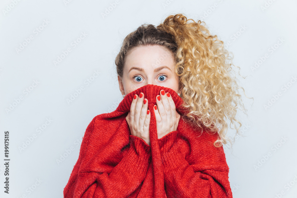 Shocked scared beautiful female stares at camera, being afraid of something awful, covers face with collar, dressed in red loose sweater, isolated over white background. Fear and fright concept Shocked scared beautiful female stares at camera, being afraid of something awful, covers face with collar, dressed in red loose sweater, isolated over white background. Fear and fright concept